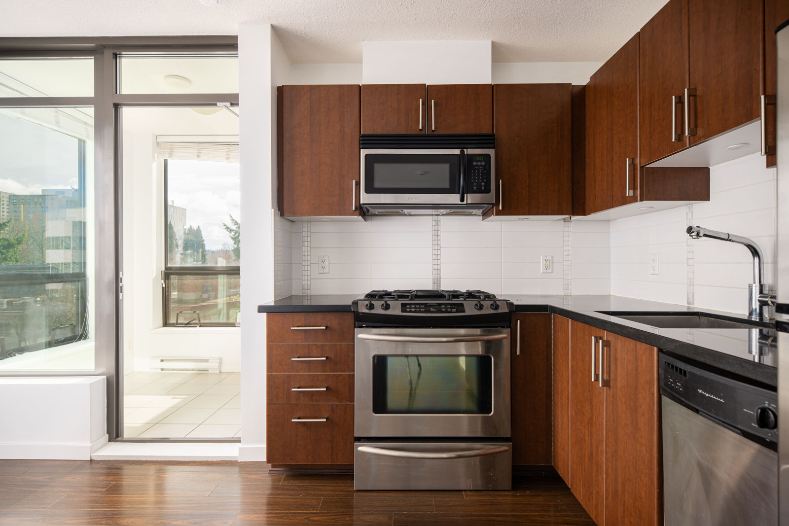 Modern kitchen with brown cabinets, stainless steel appliances, black countertops, and white tile backsplash. A glass door leads to a bright enclosed balcony.