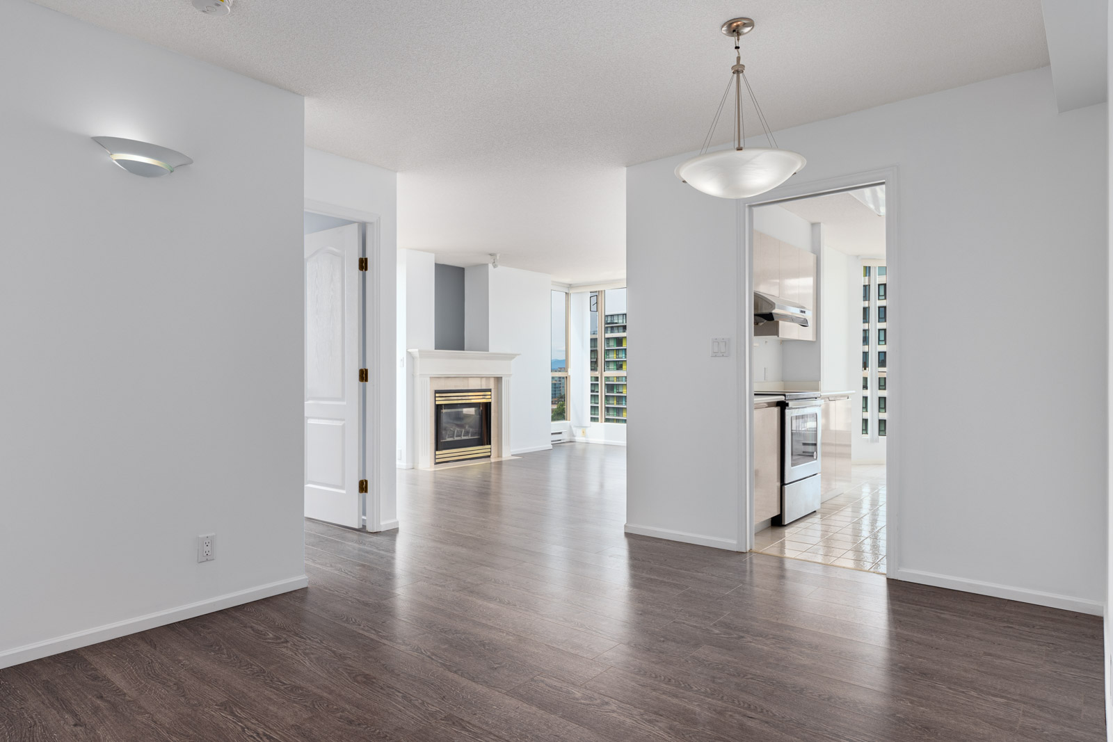 Unfurnished apartment interior with dark wood flooring, white walls, a modern light fixture, a kitchen with stainless appliances, and a living area with a fireplace and large windows.