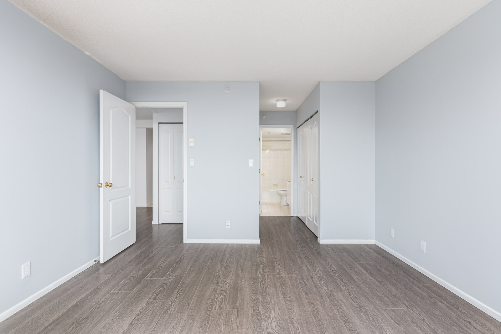 Empty room with light gray walls, dark wood flooring, white doors, and an open doorway leading to a bathroom in the background.