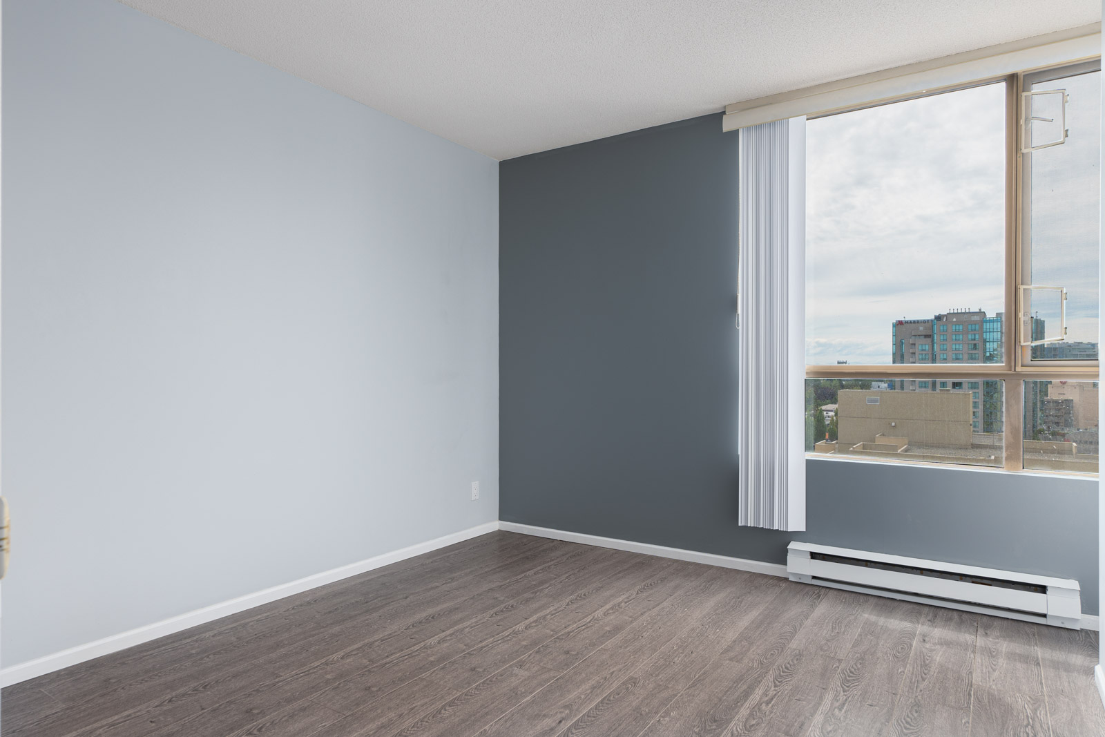Empty room with gray walls, large window with vertical blinds, and dark wood flooring. A cityscape is visible through the window under a partly cloudy sky.
