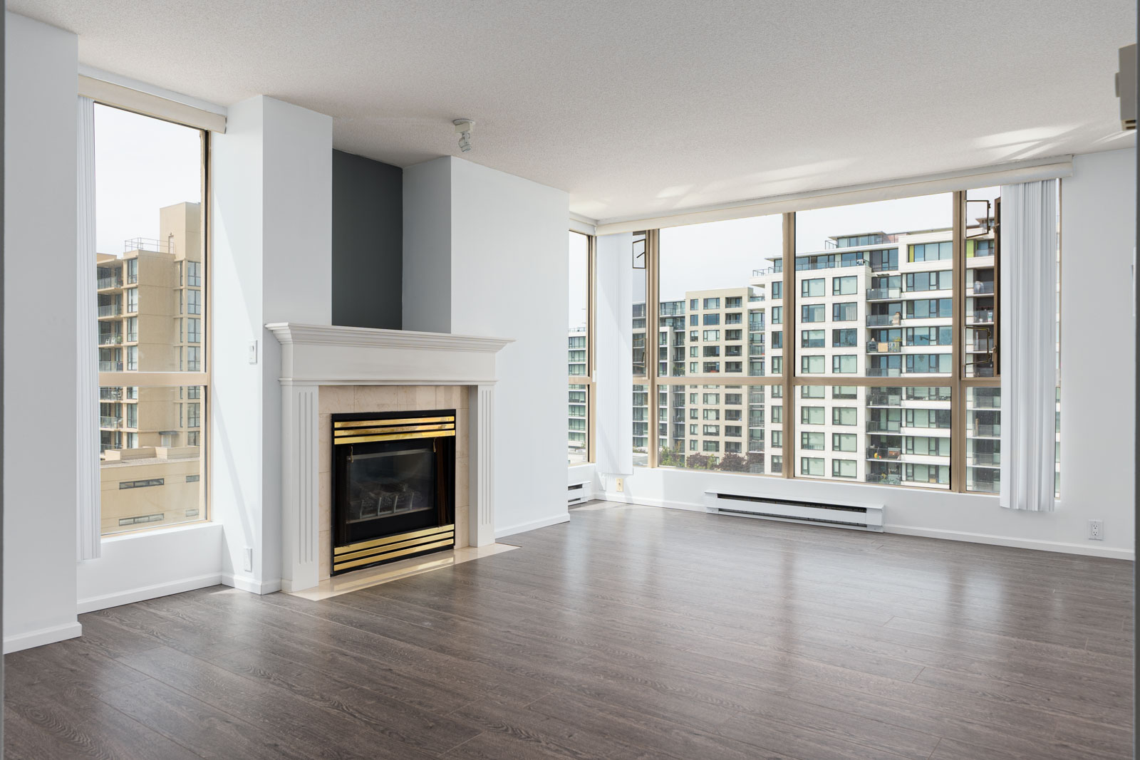 Unfurnished modern living room with large windows, wood flooring, a white fireplace with gold trim, and city buildings visible outside.