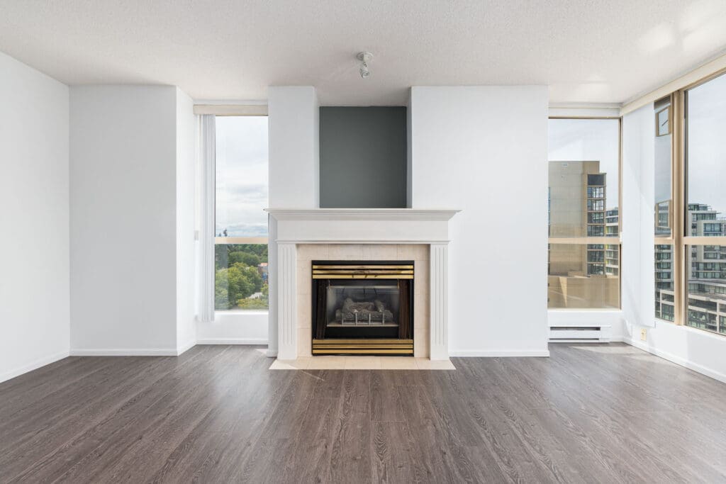 Unfurnished living room with dark wood flooring, white walls, a fireplace with a gold-trimmed mantle, and large windows offering a city view.
