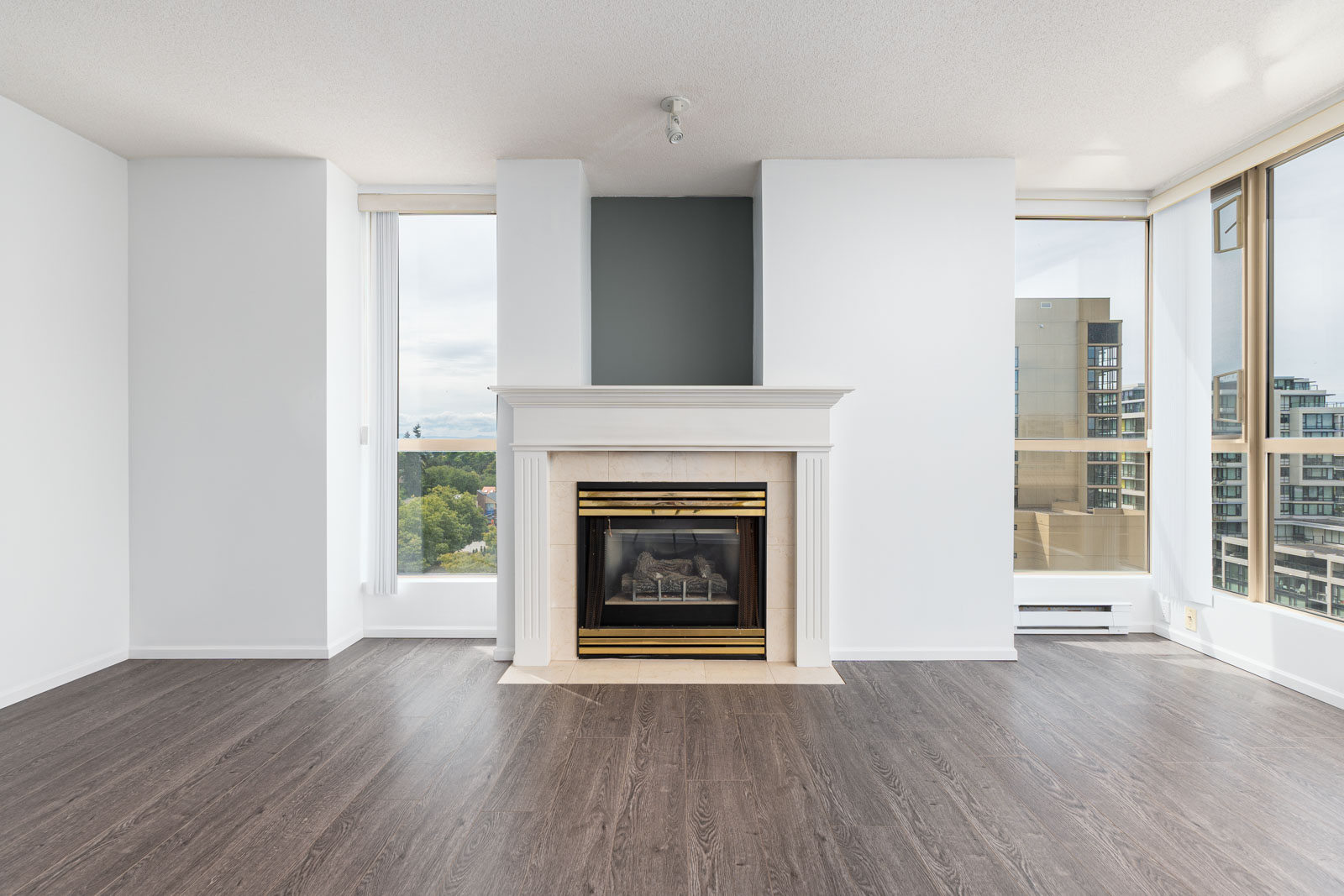 Unfurnished living room with dark wood flooring, white walls, a fireplace with a gold-trimmed mantle, and large windows offering a city view.