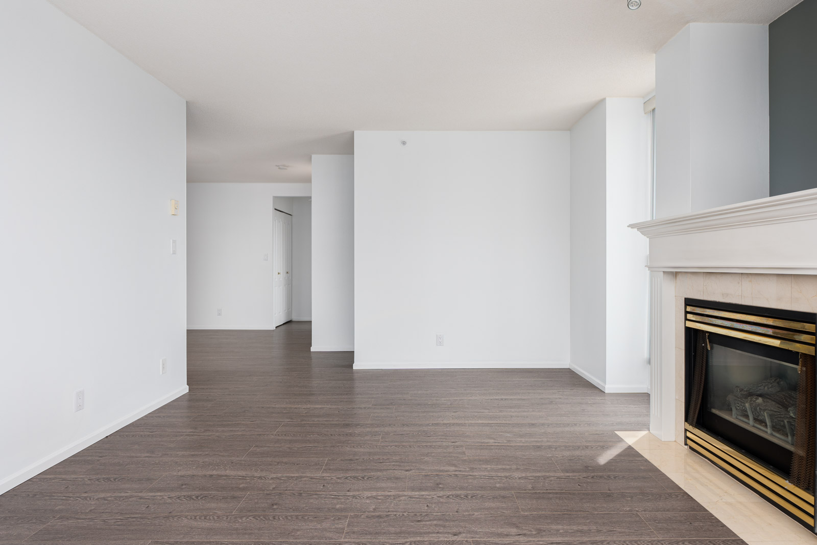 Unfurnished living room with white walls, dark wood flooring, and a white fireplace with a gold trim. Hallway leads to other rooms in the background. Natural light enters from the right.
