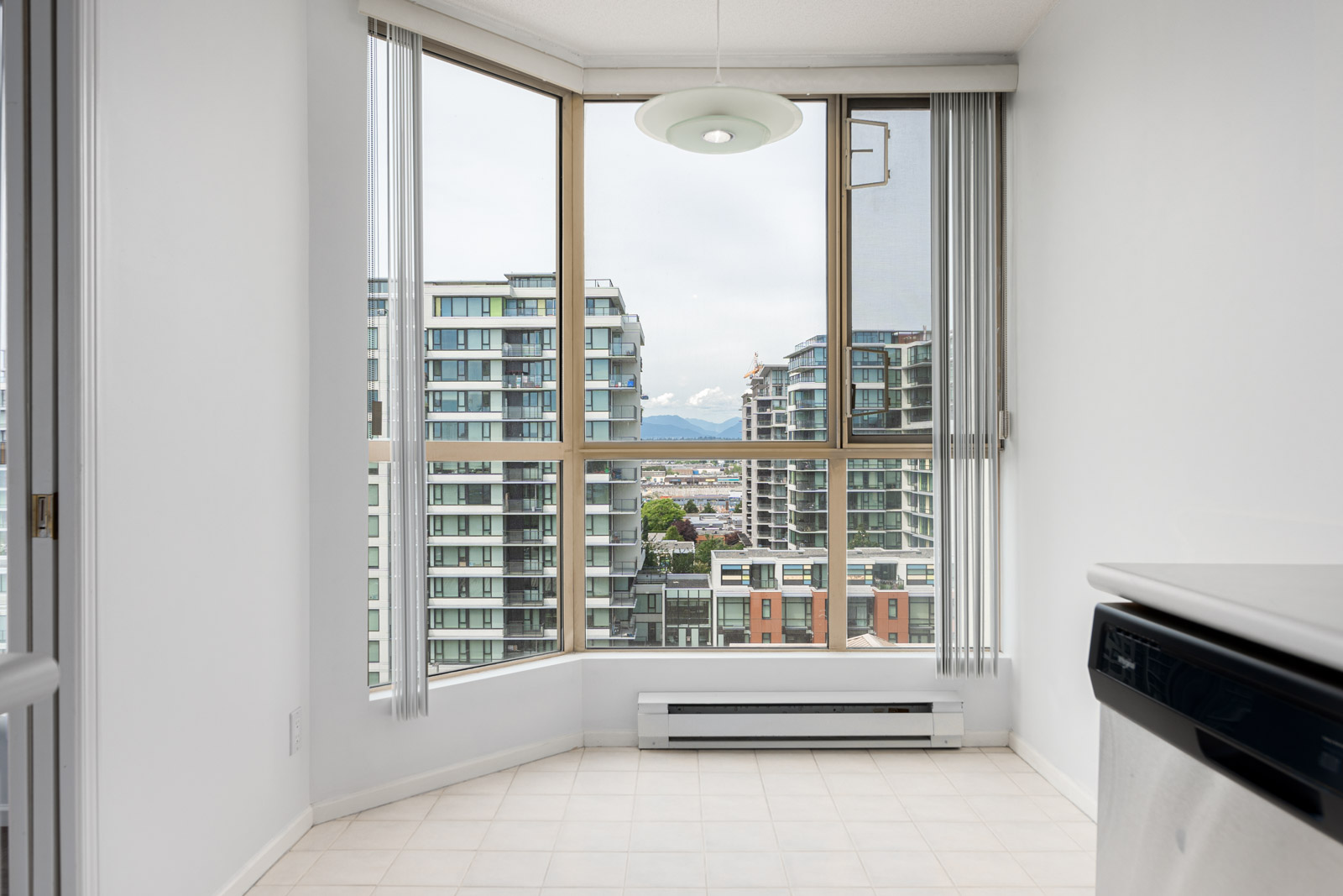 Bright room with large corner windows showing a view of modern apartment buildings and mountains in the distance, with part of a countertop and dishwasher visible.