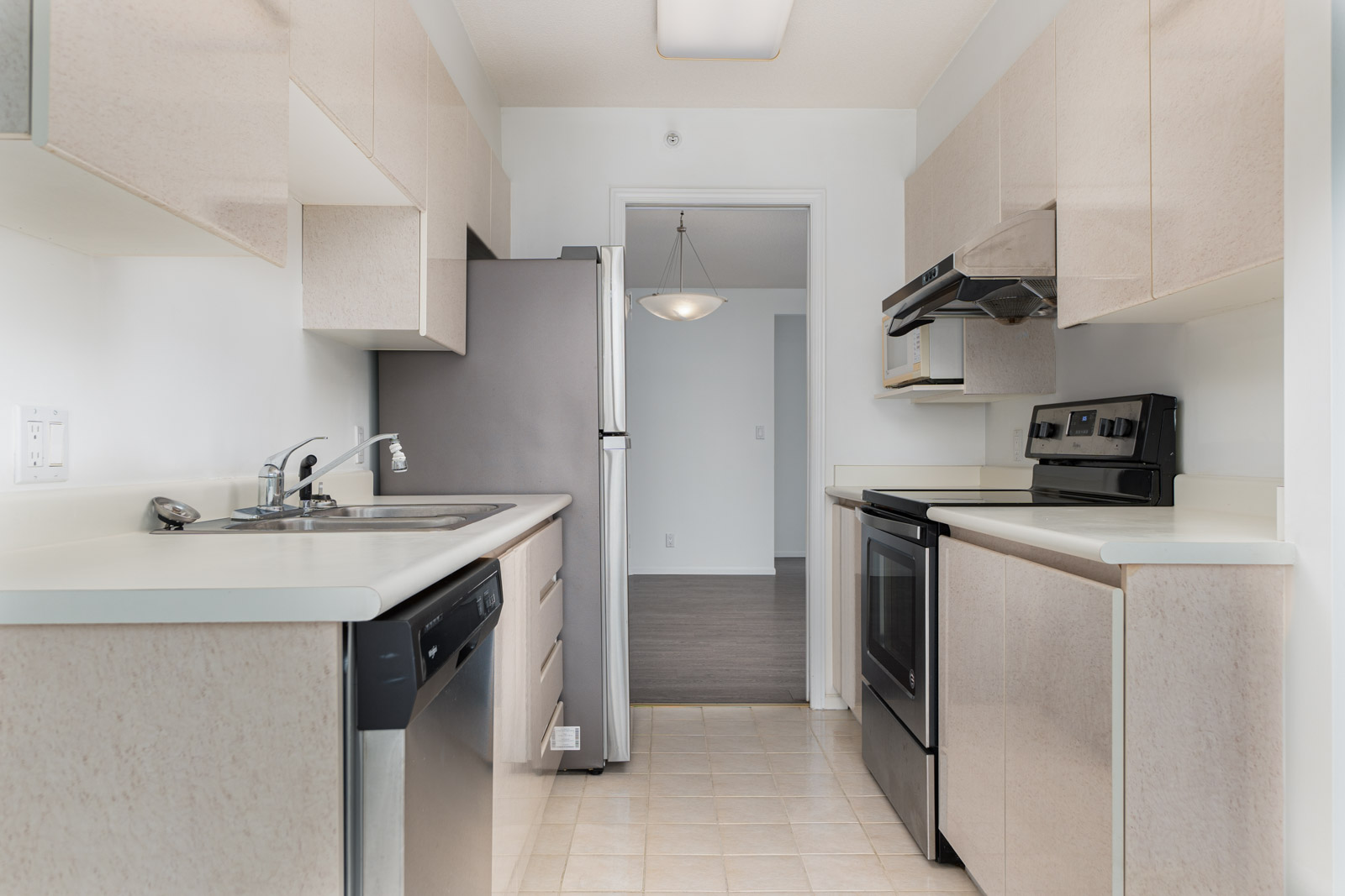 Narrow kitchen with light-colored cabinets, stainless steel appliances, electric stove, dishwasher, and sink, leading to a dining area with a hanging light fixture.