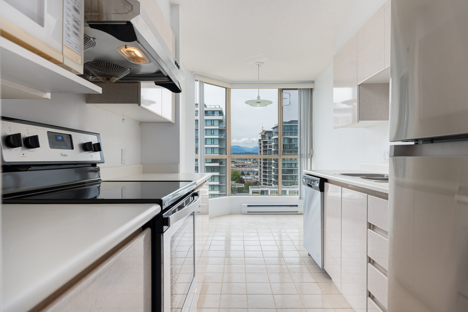 Modern kitchen with white cabinets and appliances, tiled floor, and large window offering a view of city buildings and mountains in the background.