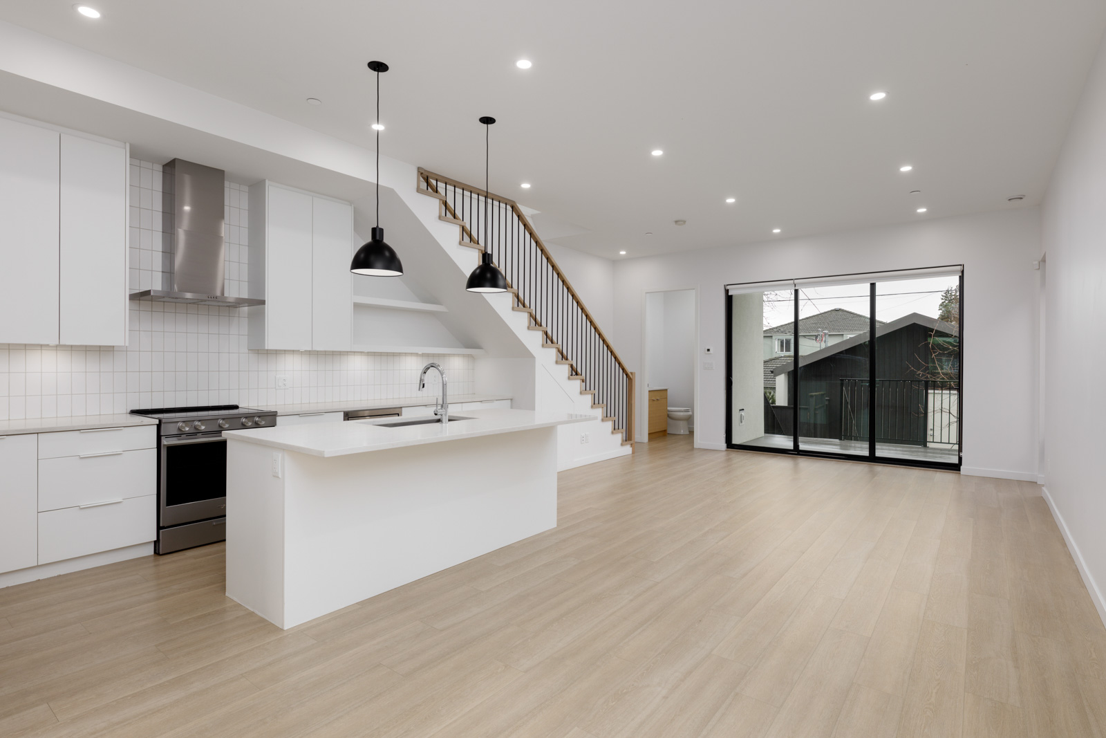Modern open-plan kitchen and living area with white cabinets, island, pendant lights, wooden floors, staircase, and large glass doors leading to a balcony.