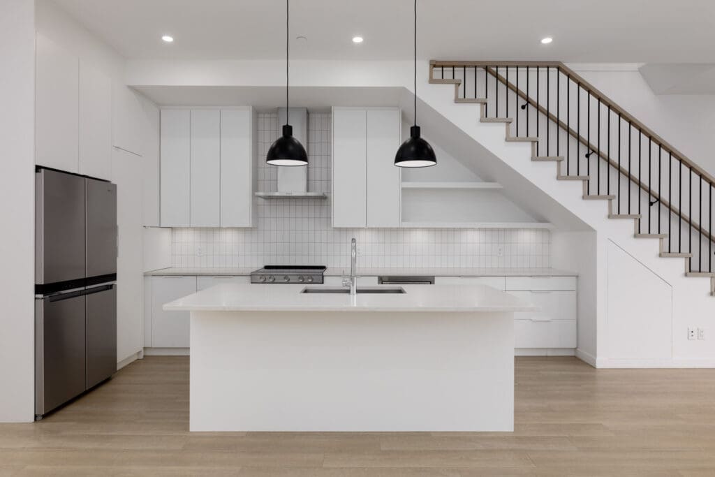 Modern kitchen with white cabinets, island countertop, stainless steel appliances, two black pendant lights, and open staircase with wooden steps.