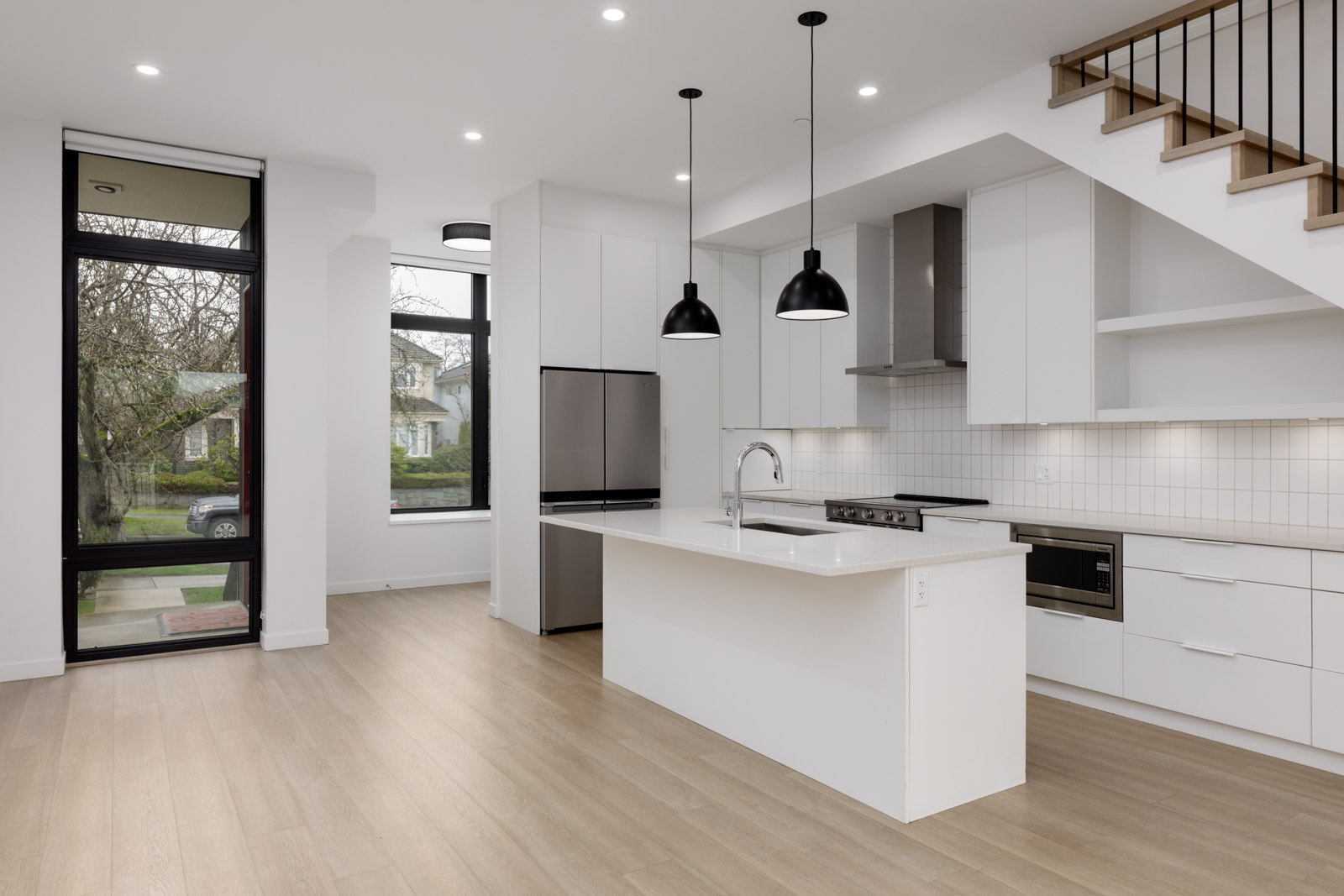 Modern white kitchen with an island, stainless steel appliances, pendant lights, wood flooring, large windows, and stairs with a metal railing.