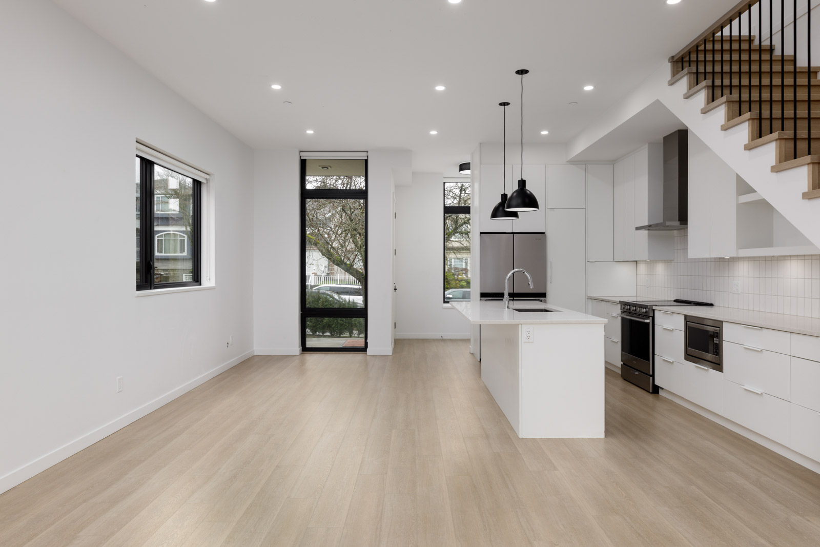 Modern, unfurnished kitchen and living area with light wood floors, white cabinets, island, pendant lights, large windows, and staircase with metal railing.