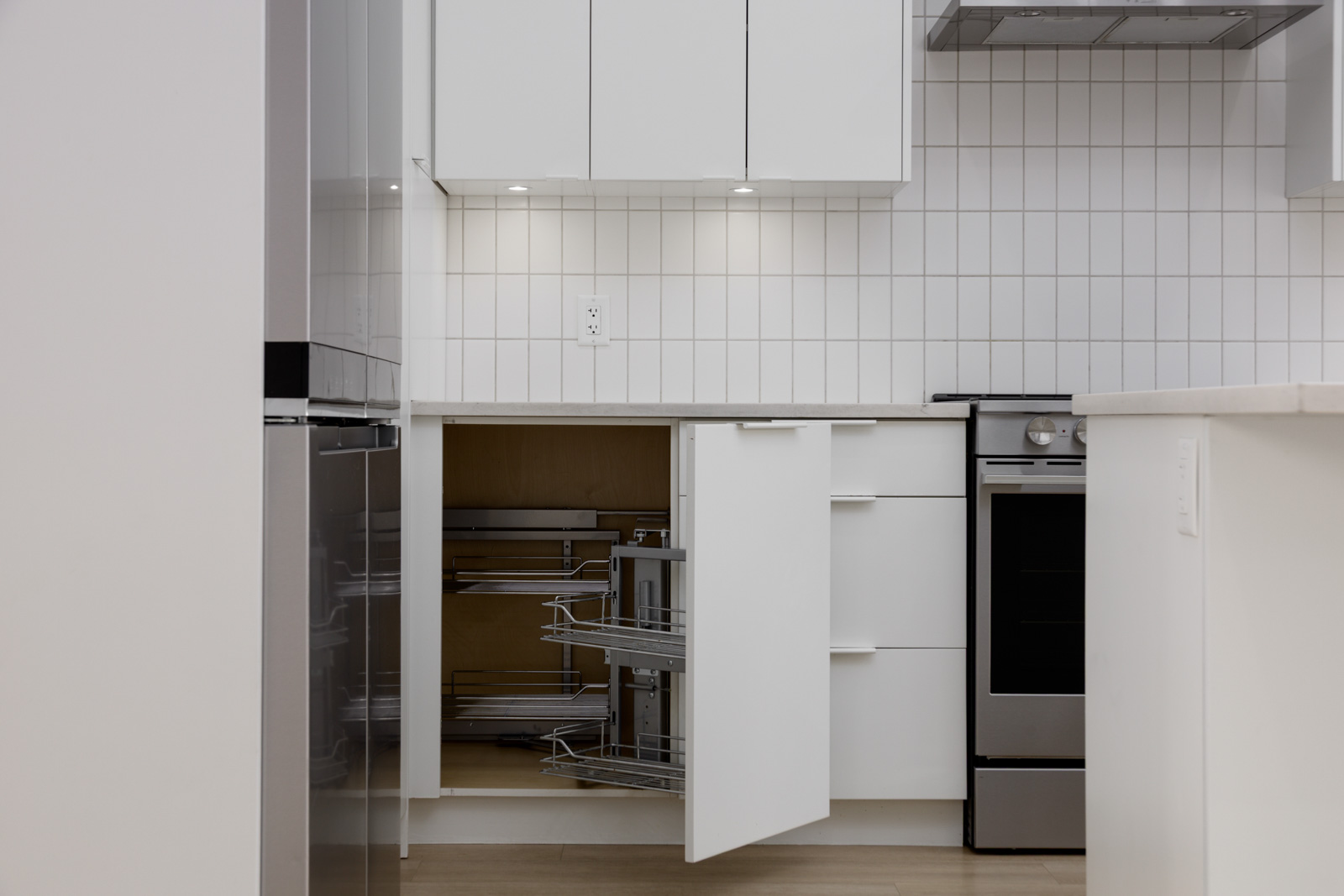Modern kitchen with white cabinets, one lower cabinet open showing pull-out metal shelves, tiled backsplash, stove, and wood flooring.