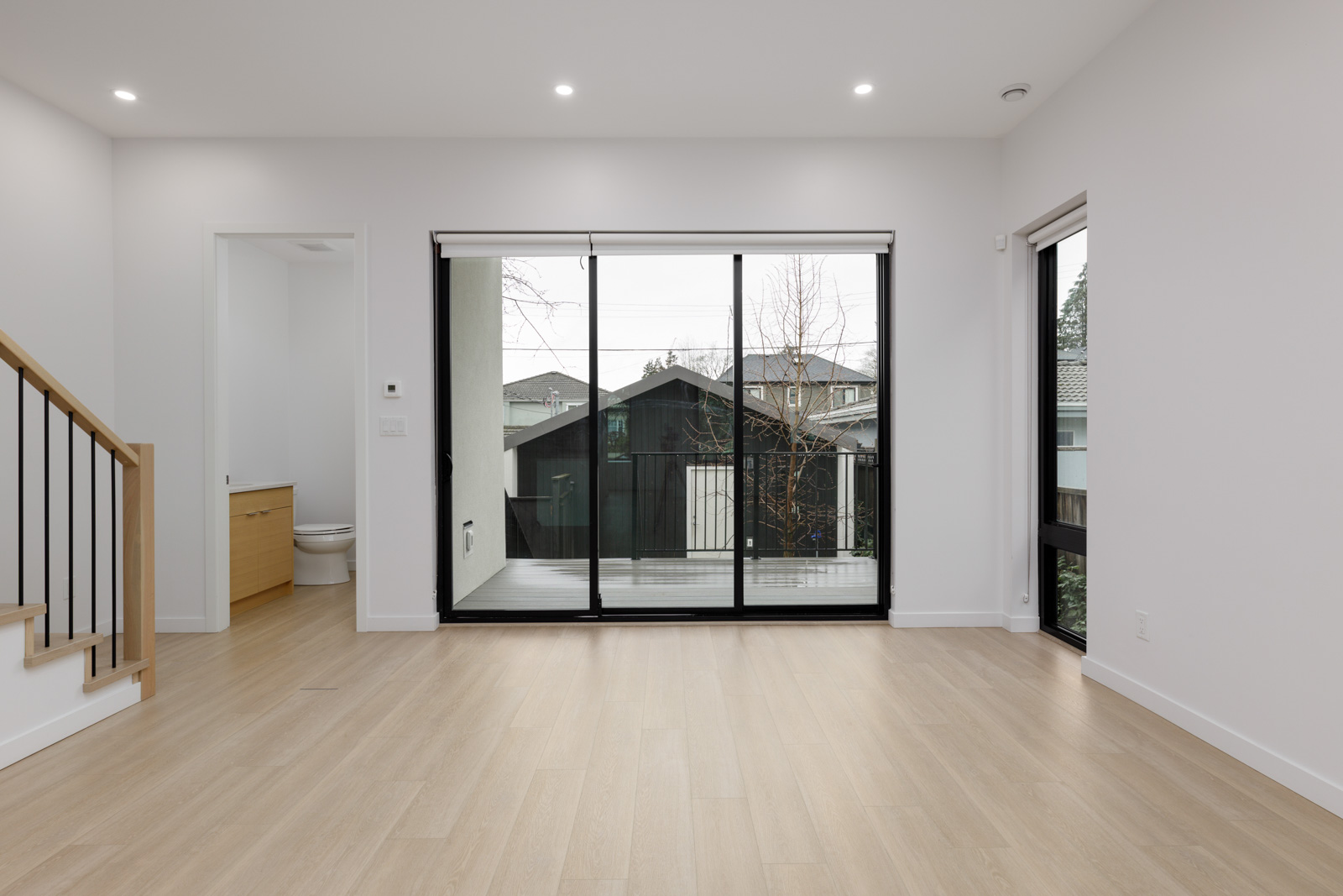 Bright, empty room with light wood flooring, large sliding glass doors opening to a balcony, a window on the right, and a wooden staircase on the left. A small bathroom is visible to the left.
