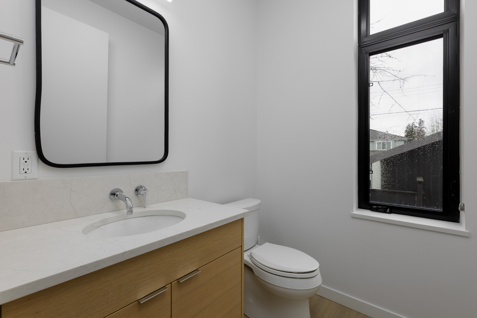 Modern bathroom with a light wood vanity, white countertop, wall-mounted faucet, rectangular mirror, toilet, and a large window with a black frame.