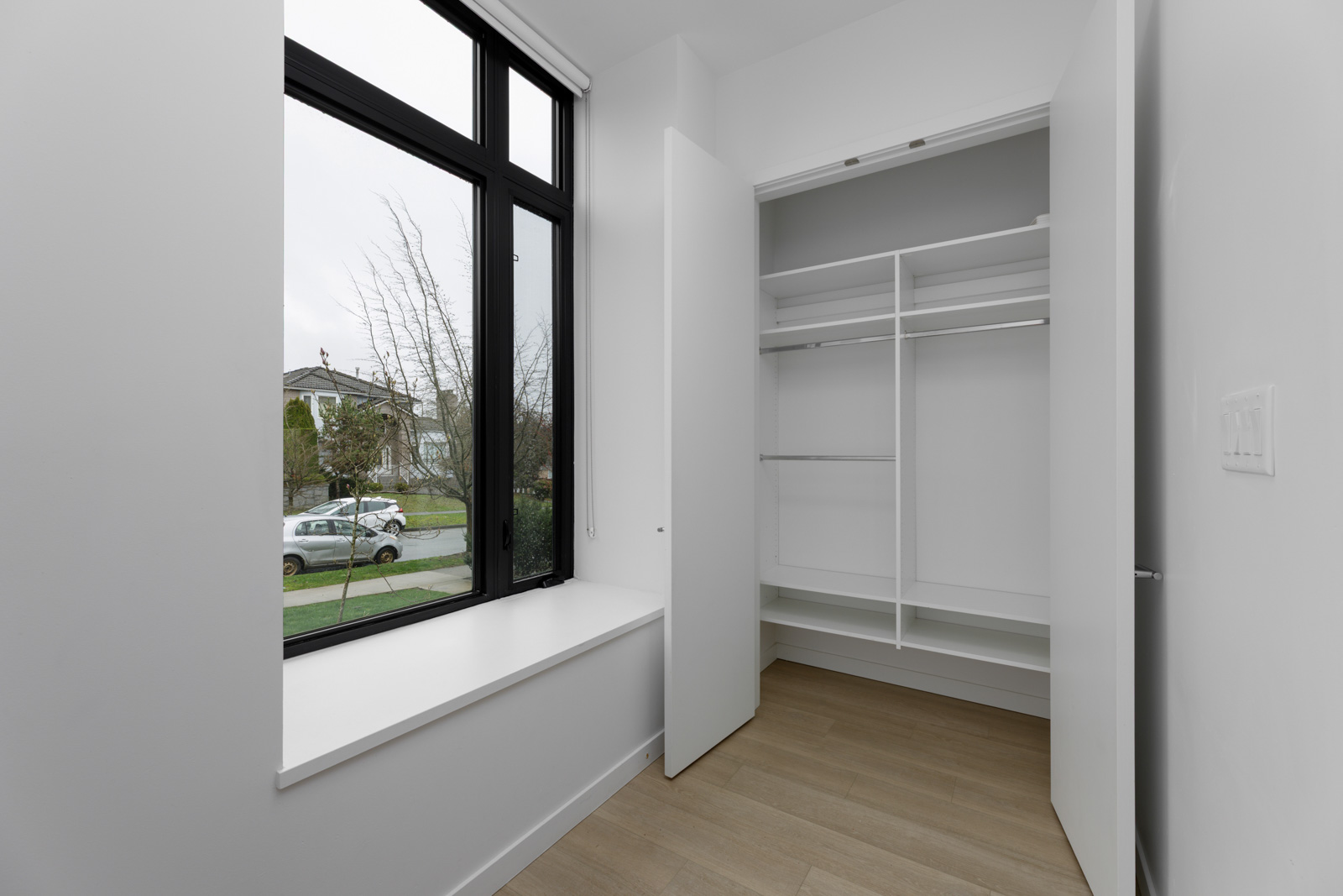 A small empty room with wood flooring, a large window with a windowsill, and an open built-in closet with shelves and hanging rods.