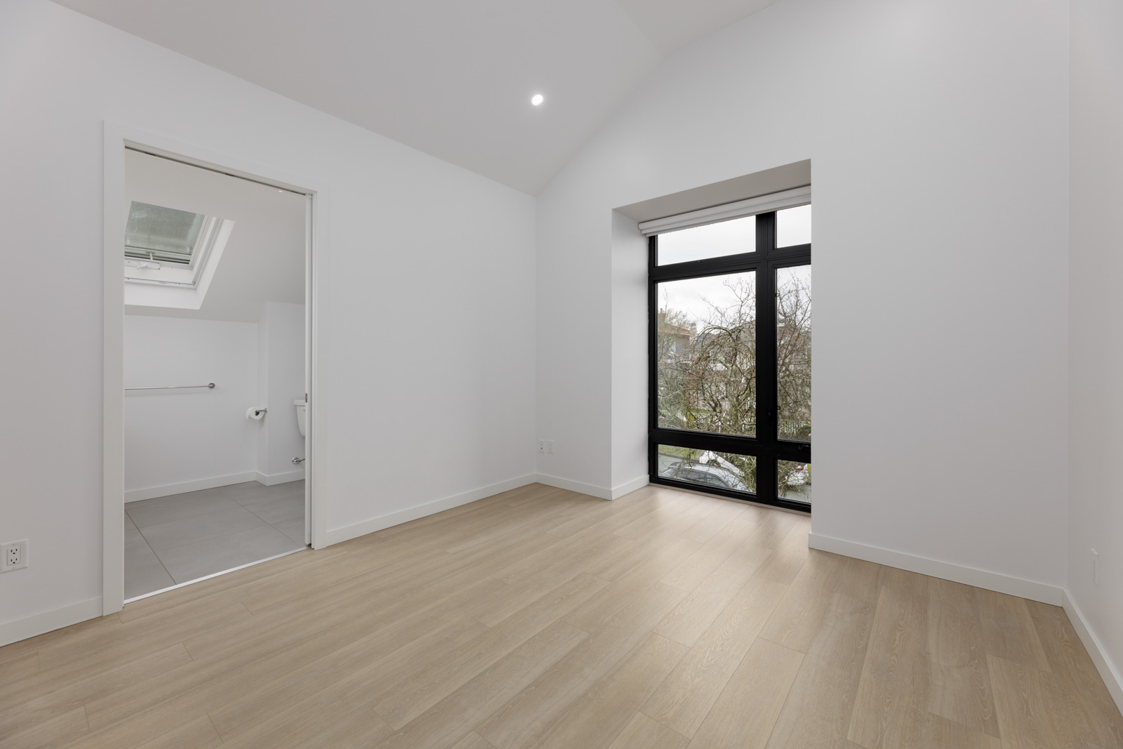Empty room with light wood flooring, white walls, an open doorway leading to a bathroom, and a large window with a view of trees outside.