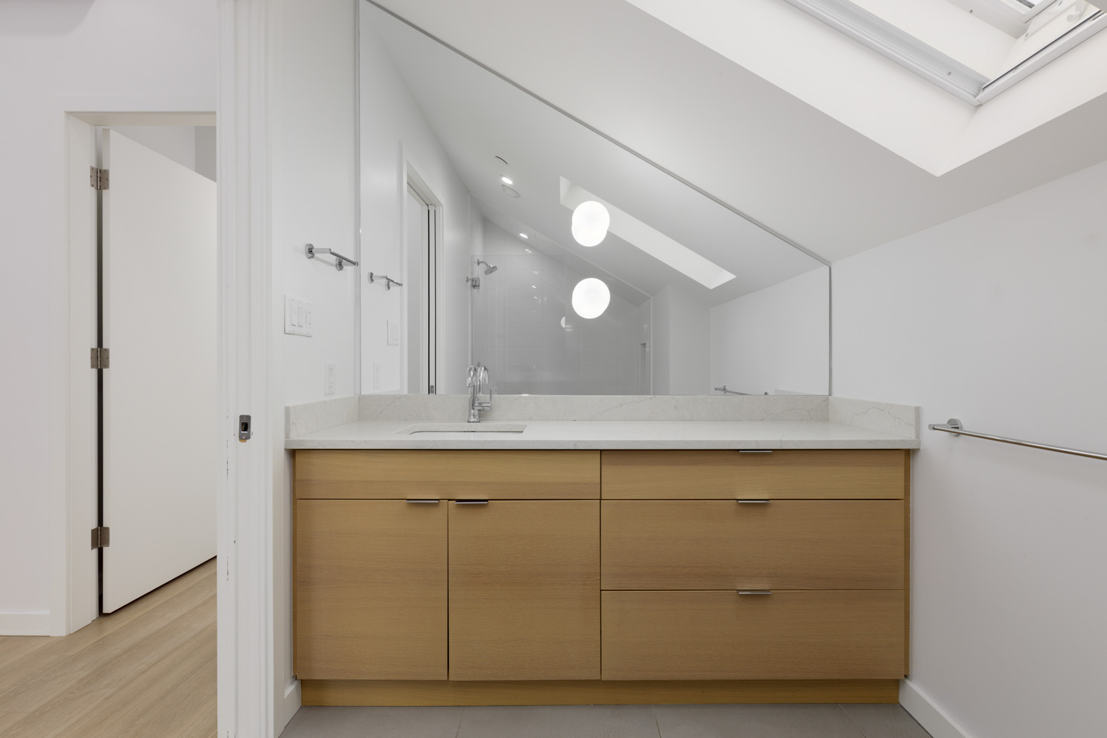 Modern bathroom with a light wood vanity, white countertop, large mirror, and angled ceiling with two round light fixtures. A door is open to an adjacent room.