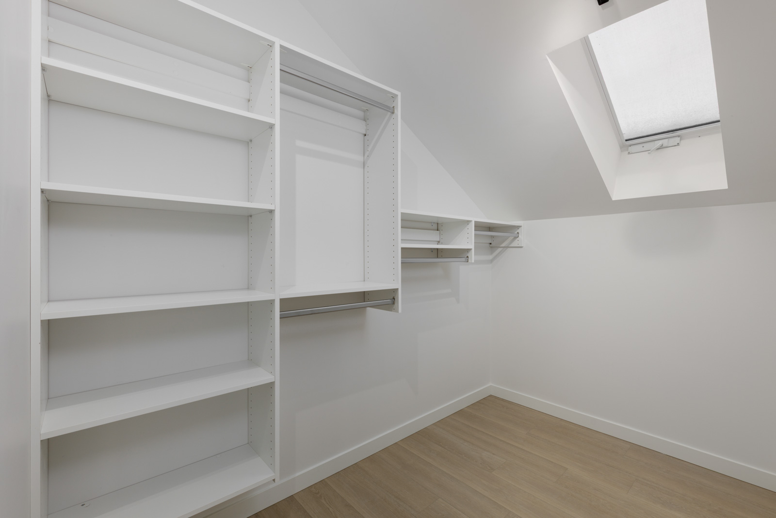Empty walk-in closet with white shelves and hanging rods, light wood floor, slanted ceiling, and a skylight window letting in natural light.