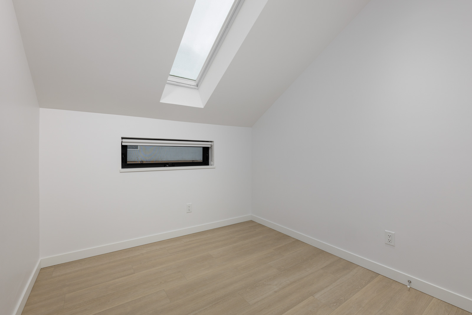Small, empty room with light wood flooring, white walls, a rectangular side window with a shade, and a skylight on the sloped ceiling.