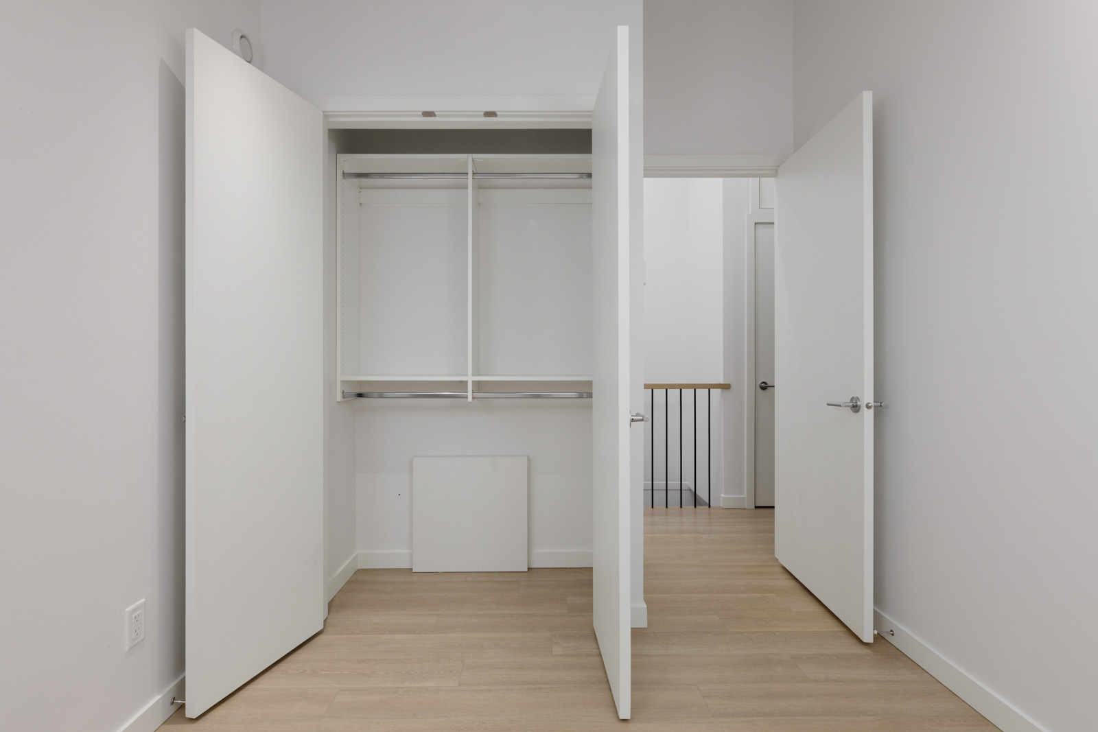 Empty closet with open double doors, a hanging rod, and shelf inside a room with light wood flooring and white walls; adjacent door opens to hallway.