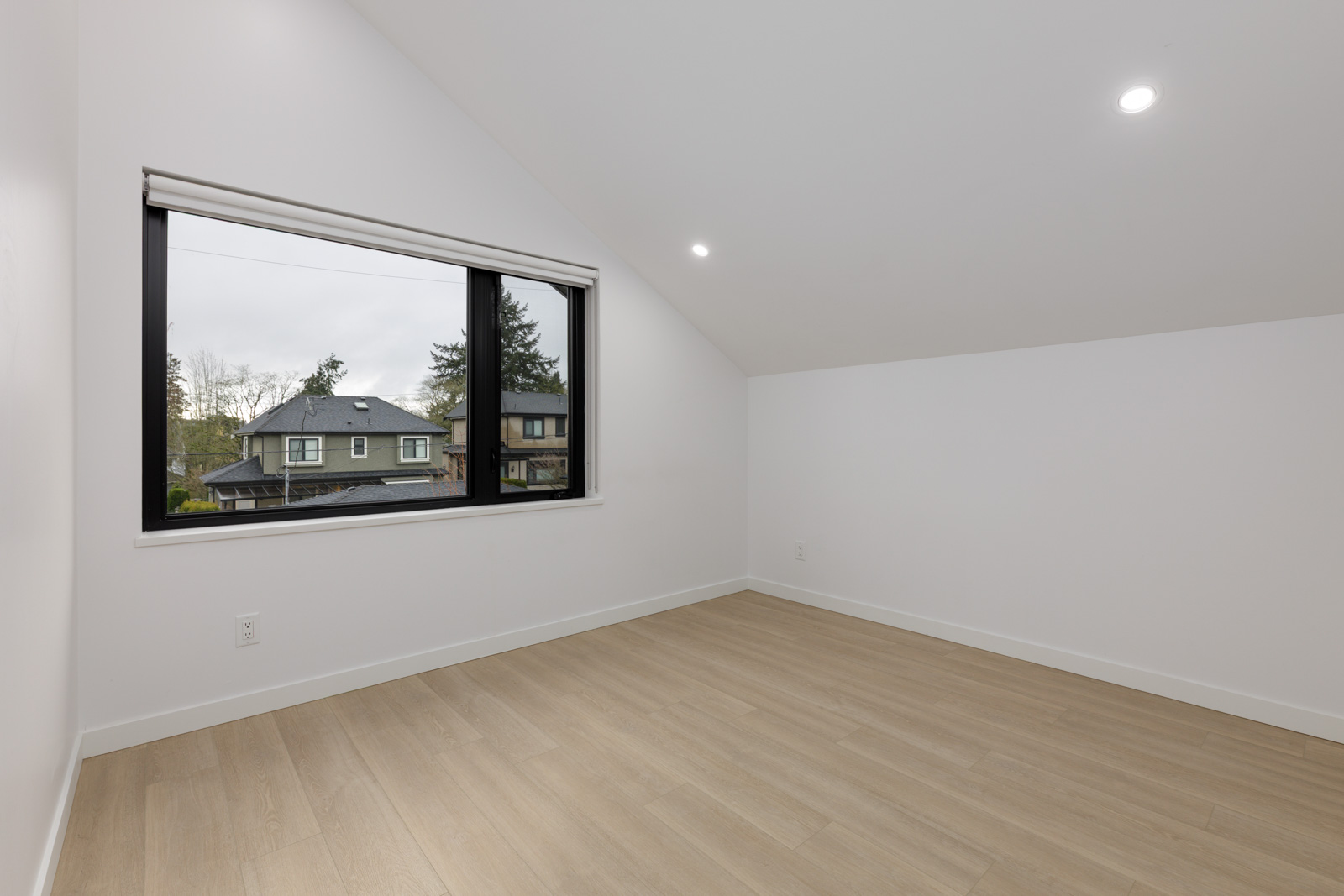 Empty room with light wood flooring, white walls, a large window showing neighboring houses, and recessed ceiling lights.
