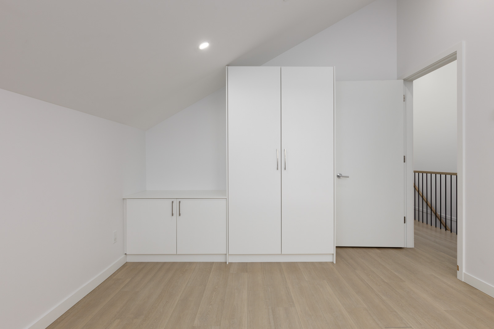 A minimalist room with light wood flooring, white walls, a wardrobe, a cabinet, and an open door leading to a hallway with a metal railing.