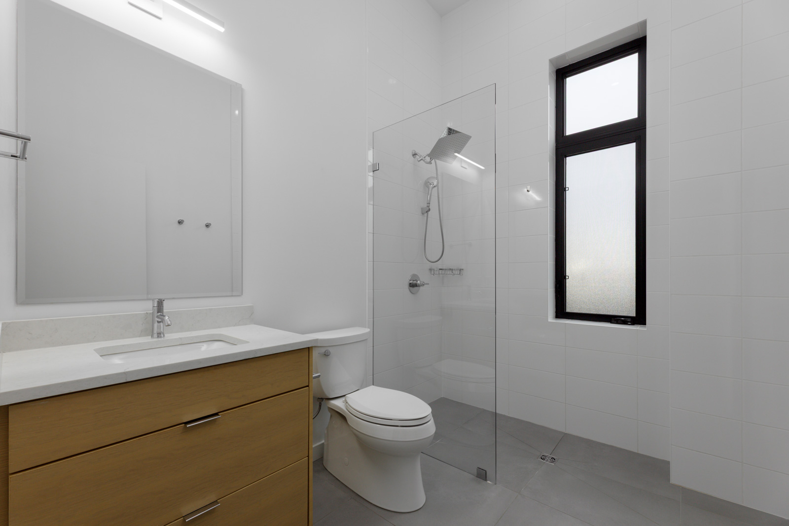 Modern bathroom with a wooden vanity, white sink, wall mirror, toilet, and a glass-enclosed shower next to a tall frosted window.