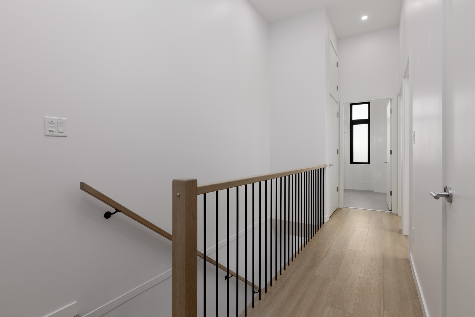 A modern hallway with light wood flooring, white walls, a black metal and wooden handrail on the left, and doors leading to adjoining rooms.