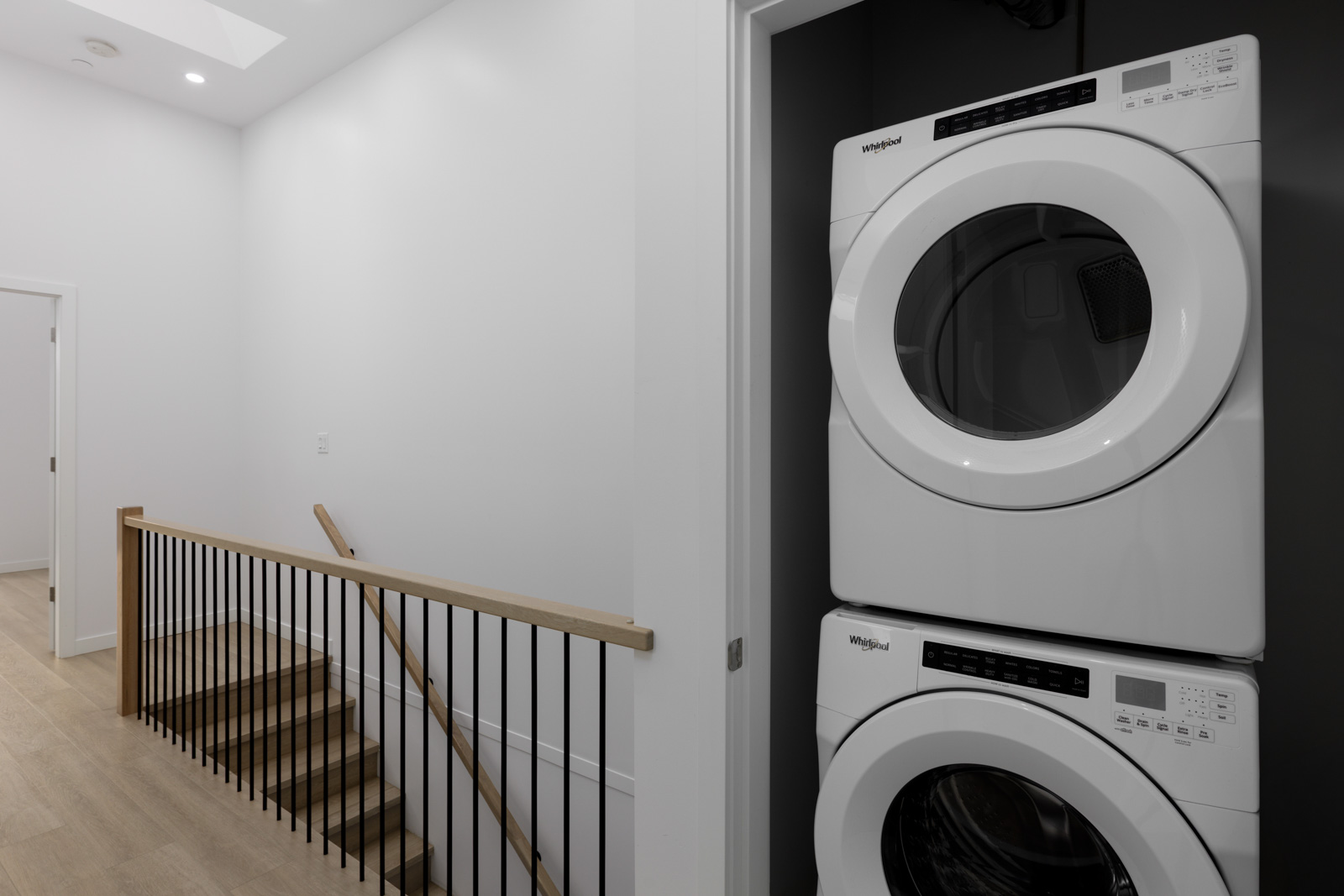 A stacked Whirlpool washer and dryer are placed in a recessed laundry area next to a hallway with wooden stairs and a railing.