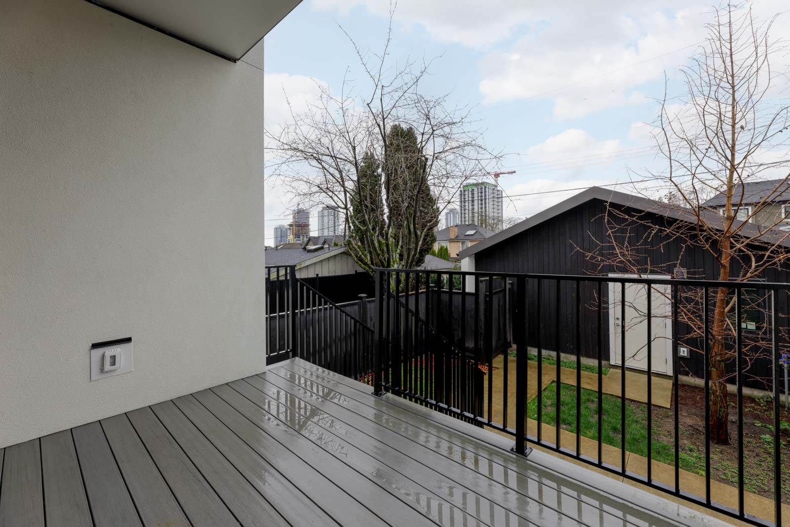 A modern balcony with gray decking and black railing overlooks a backyard with trees, lawn, and neighboring buildings under a cloudy sky.