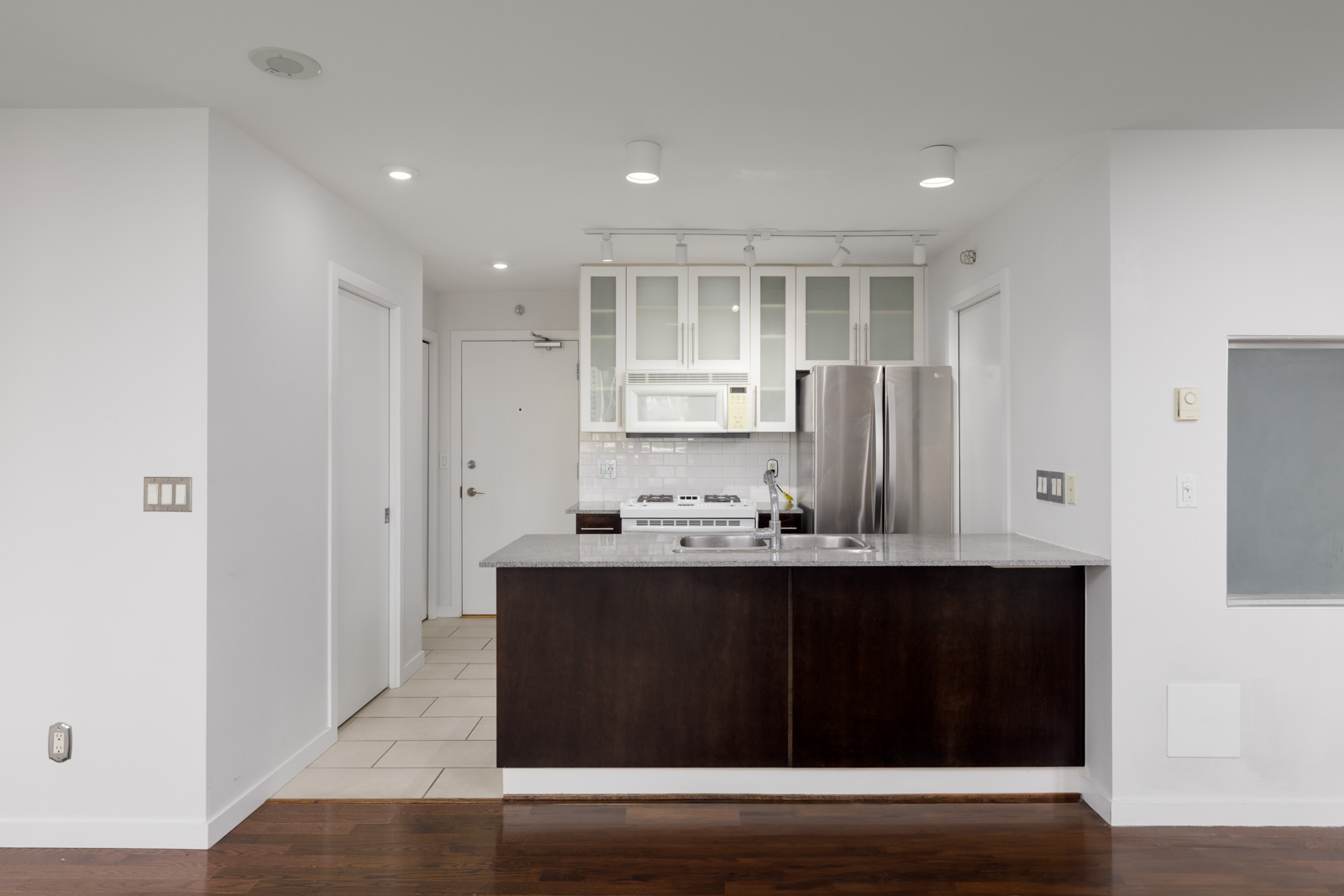 Modern kitchen with stainless steel appliances, dark wood lower cabinets, white upper cabinets with glass doors, and a gray countertop; white walls and tile flooring.