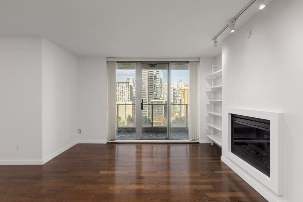 Modern empty living room with wood floors, white walls, a built-in fireplace, shelving, and glass doors leading to a balcony with a city view.
