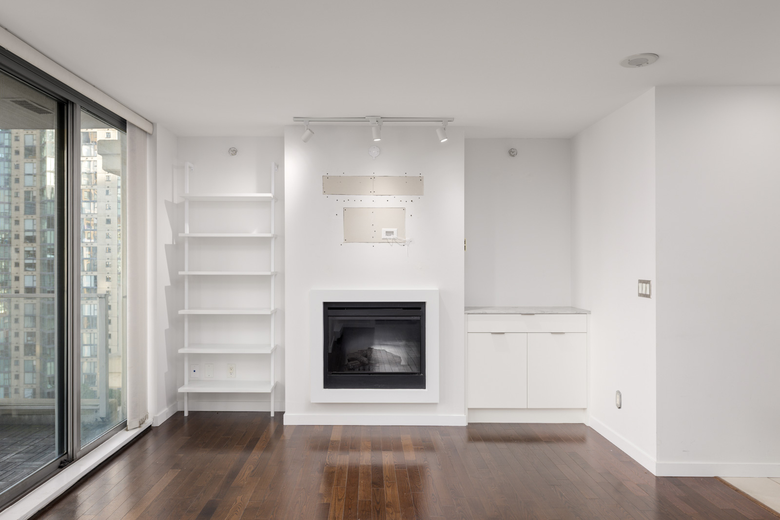 Modern unfurnished living room with hardwood floors, a fireplace, built-in shelves, white cabinets, and large windows overlooking city buildings.
