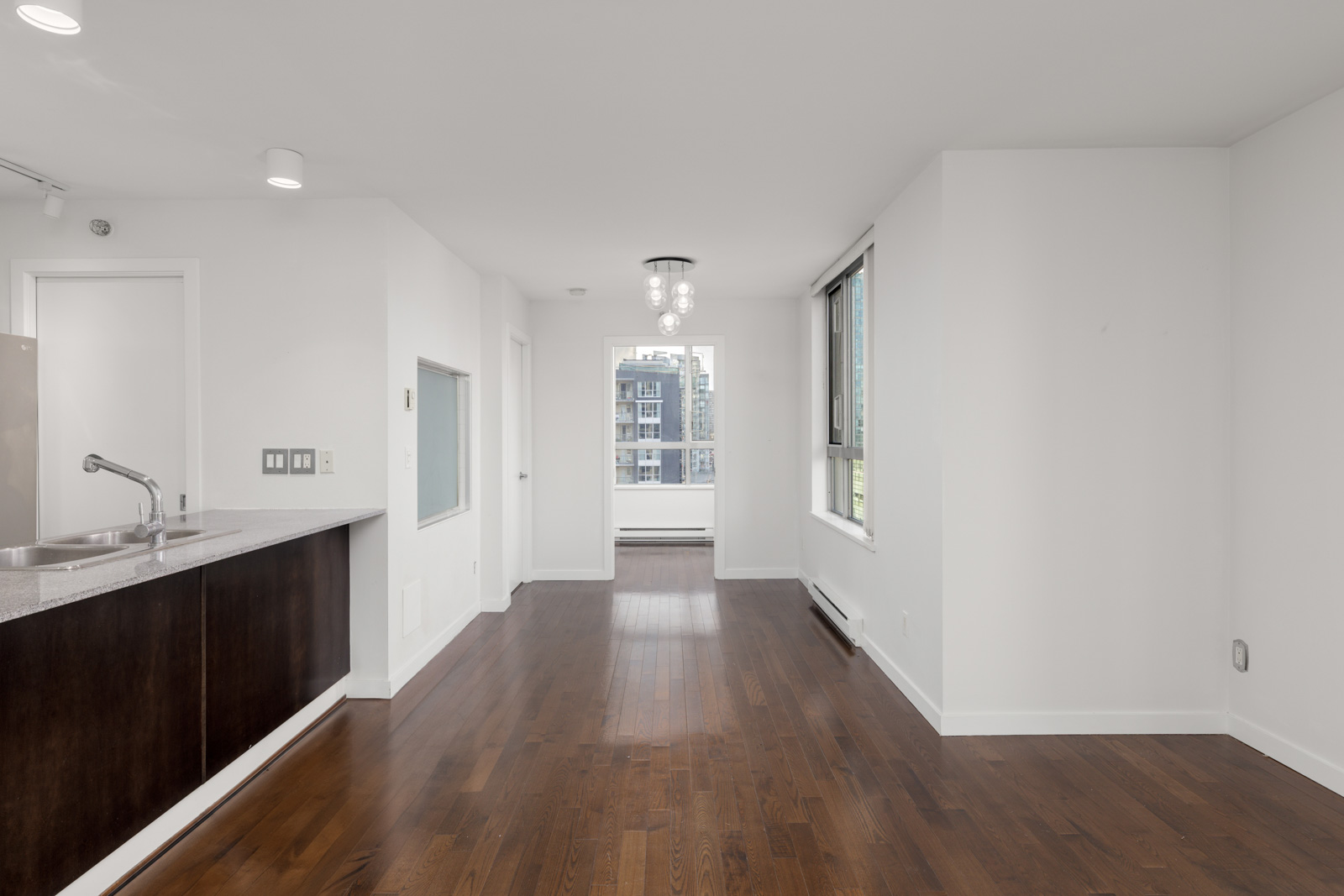 Modern, unfurnished apartment interior with wooden floors, white walls, open kitchen area, large window, and city view through the far window.