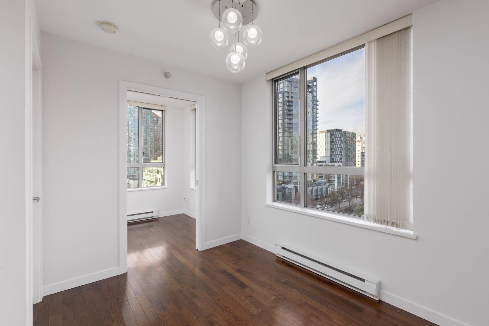 Unfurnished room with white walls, hardwood floors, large windows, city view, and modern ceiling light fixture. Adjacent room visible through open sliding doors.