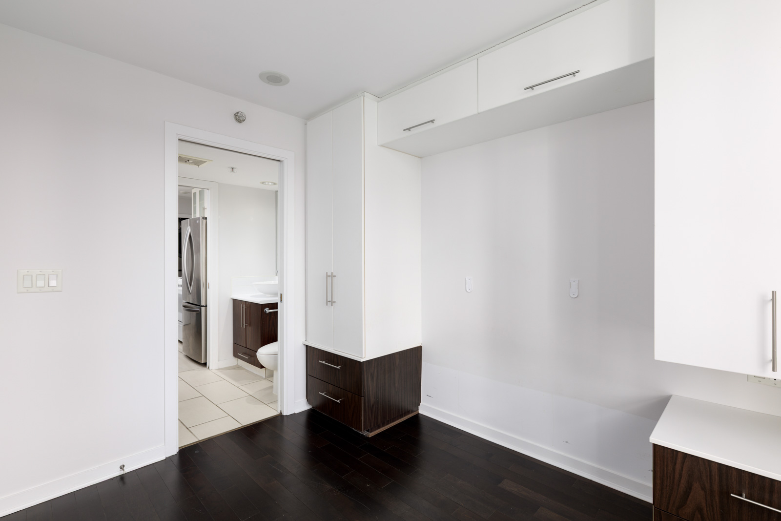 A modern, empty room with white walls, dark wood flooring, built-in cabinets, and an open doorway leading to a bathroom with tiled floors.