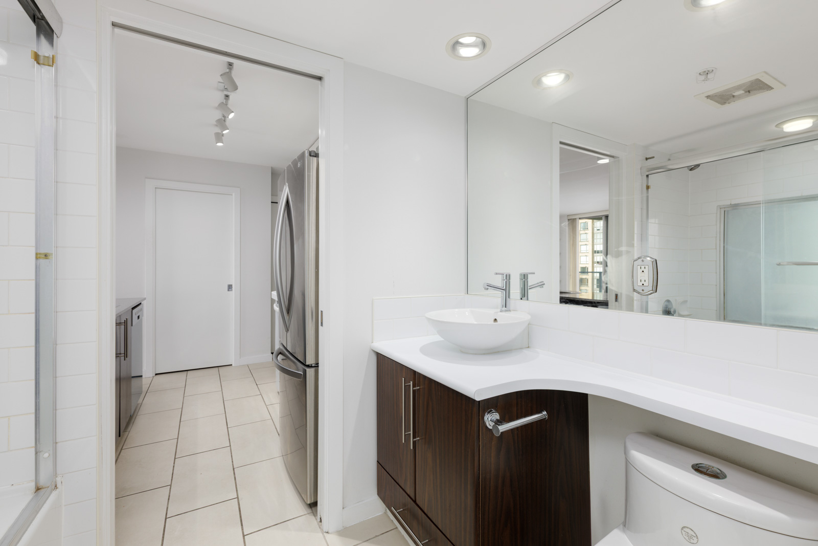 Modern bathroom with white walls, a large mirror, vessel sink on a dark vanity, toilet, and a view into a kitchen area with stainless steel refrigerator.