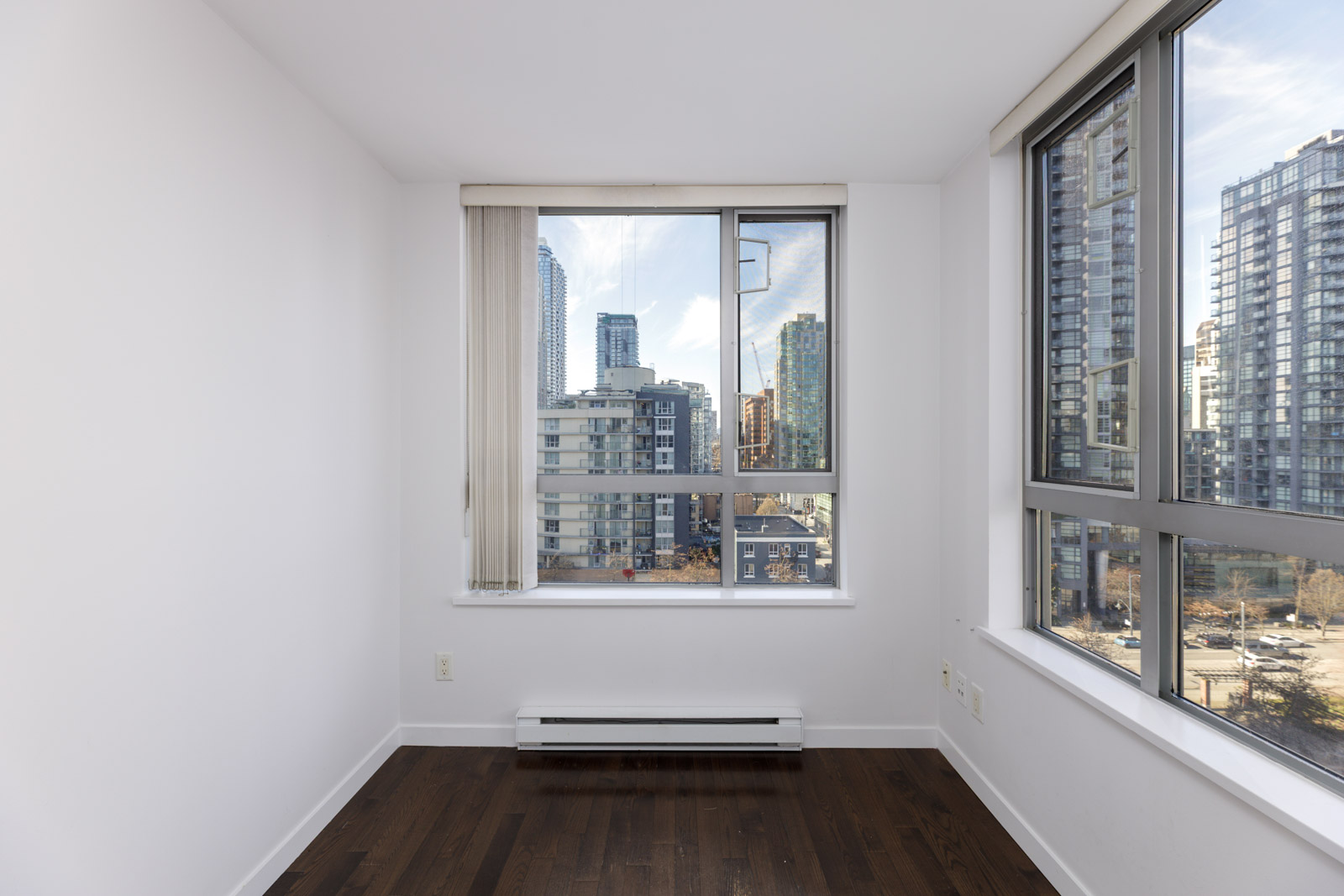 Empty room with white walls, dark wood floor, and large windows showing a cityscape with modern high-rise buildings under a partly cloudy sky.