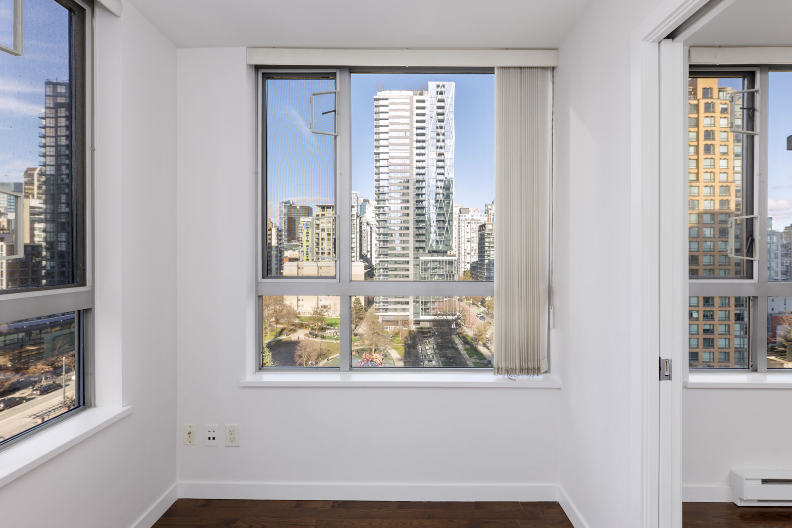 A bright, empty room with white walls, large windows, and a view of modern city buildings outside.