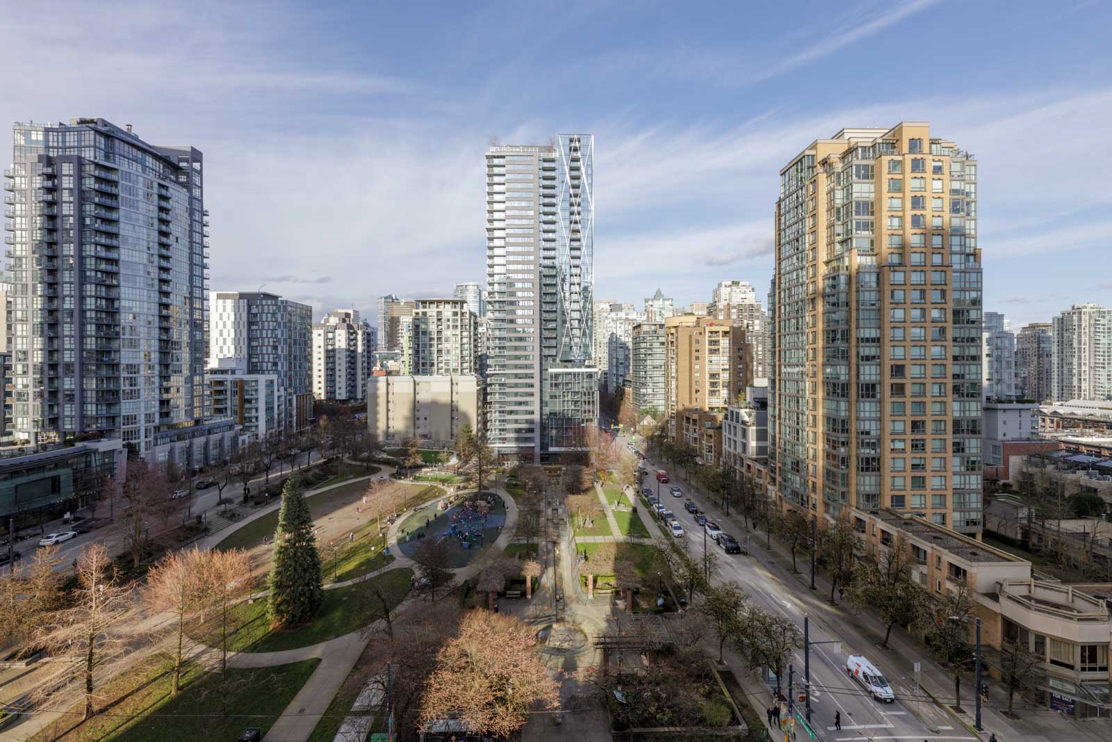 View of a city park surrounded by high-rise residential buildings under a partly cloudy sky, with streets and cars visible along the park’s borders.