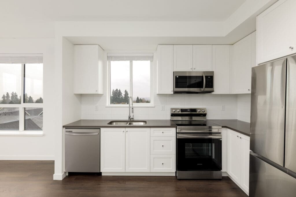 Modern kitchen with white cabinets, stainless steel appliances, a window above the sink, and dark countertops. The floor is dark wood, and the space appears clean and bright.
