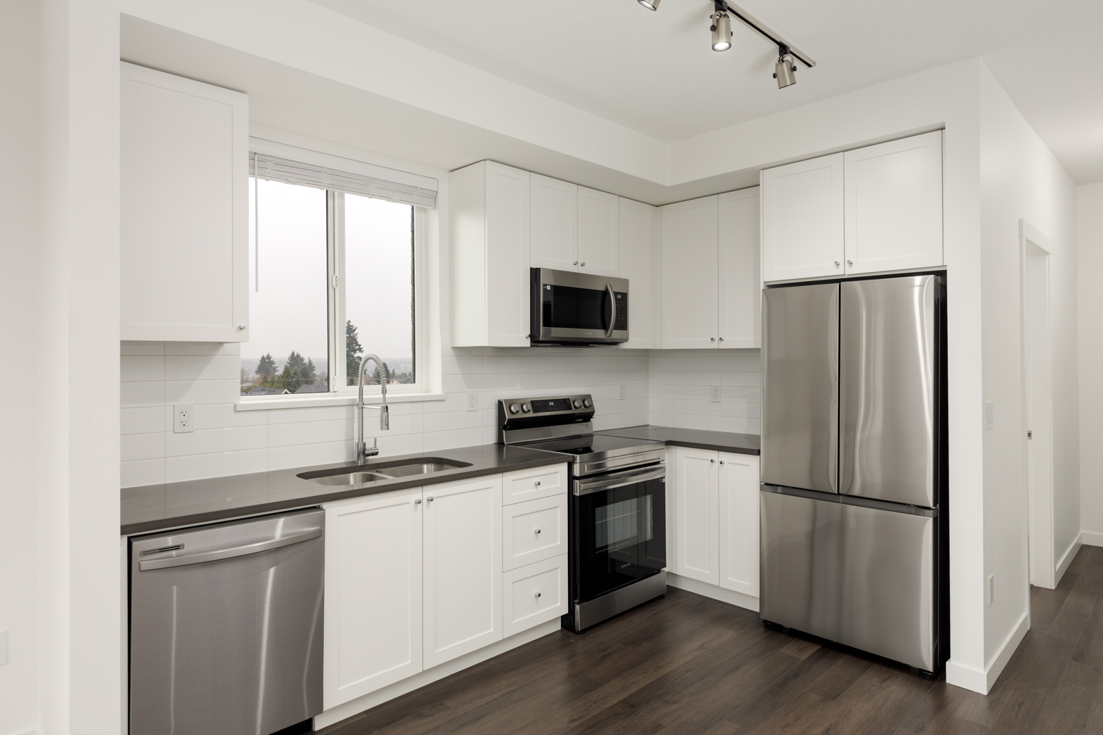 Modern kitchen with white cabinets, stainless steel appliances, dark countertops, a single window above the sink, and dark wood flooring.