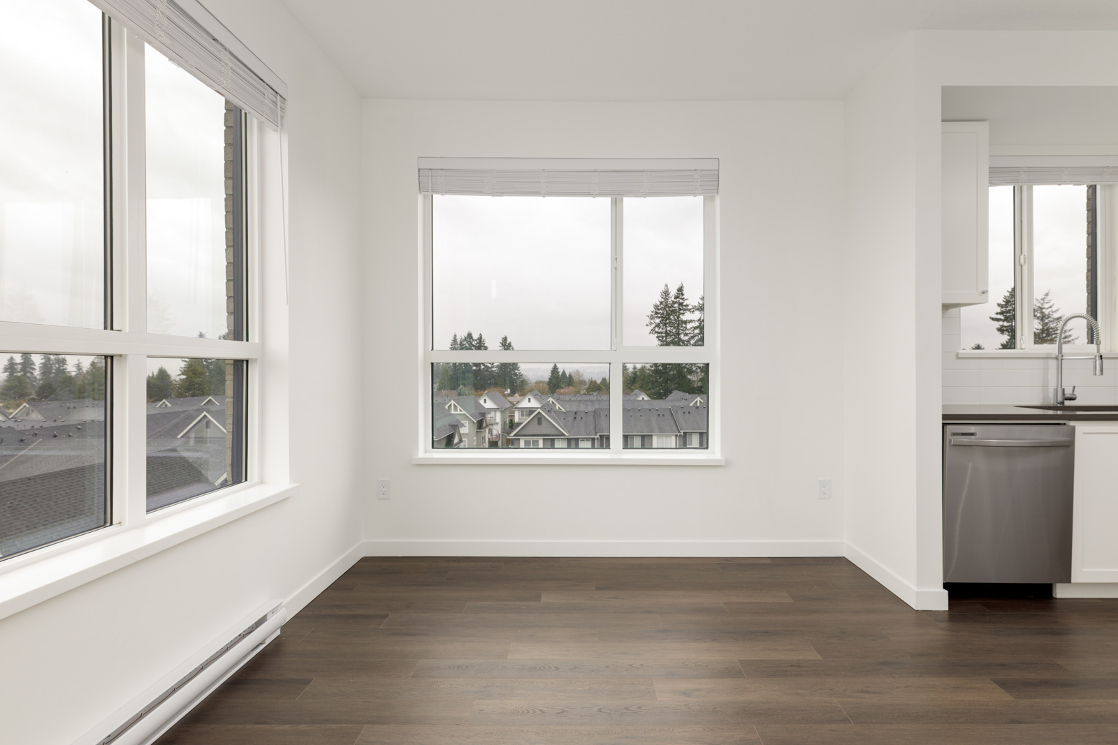 Unfurnished room with large windows, white walls, dark wood flooring, and a partial view of a kitchen area with a dishwasher and sink.
