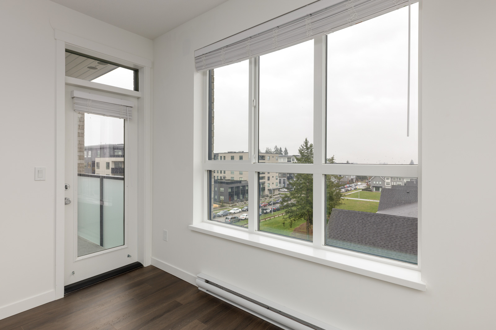Corner of a modern room with large windows, white walls, a glass door to a balcony, and dark wood flooring, overlooking buildings and a parking lot outside.