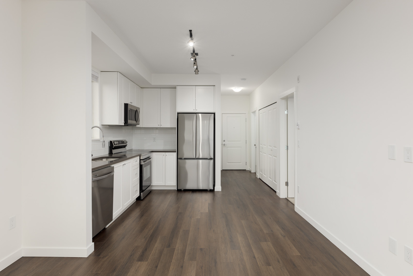 Modern kitchen with stainless steel appliances, white cabinets, and wood flooring, opening into an empty living area with white walls and closet doors.