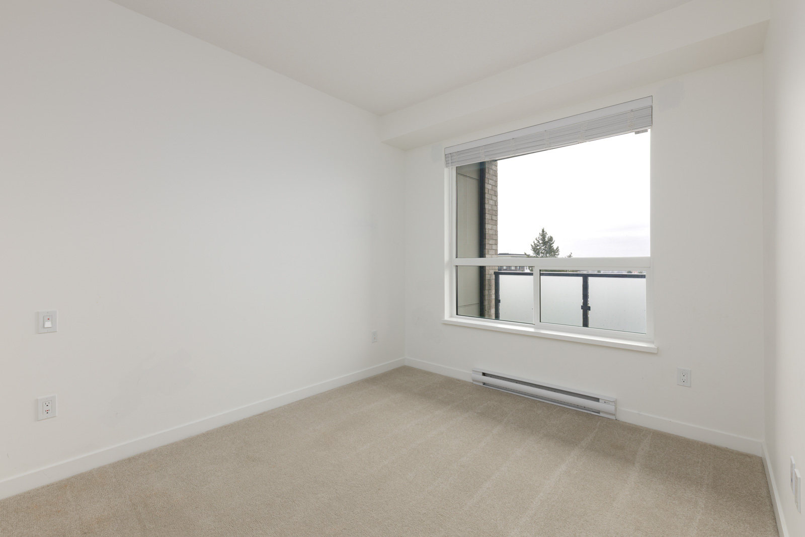 Empty room with beige carpet, white walls, a large window with blinds, and a view of a balcony and sky outside.