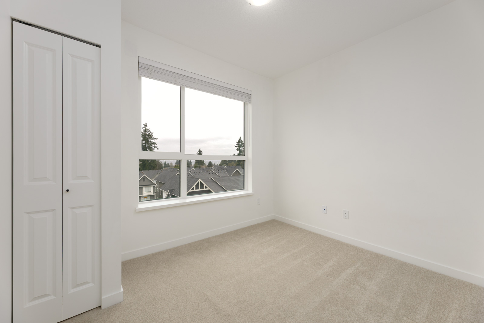 Empty room with beige carpet, white walls, a window showing rooftops and trees outside, and a closed white closet door on the left.