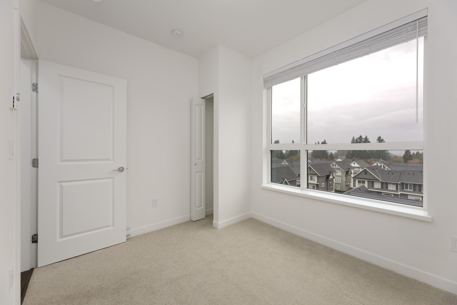 Empty, carpeted room with white walls, a large window overlooking houses and trees, and two white doors—one open and one closed.