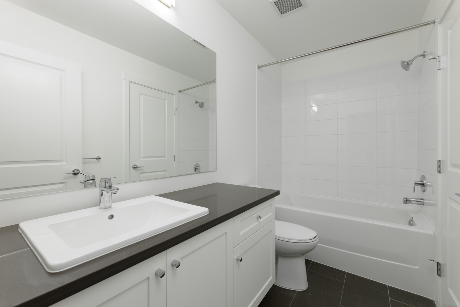 Modern bathroom with a white bathtub and shower, a toilet, a wide mirror, a black countertop, and white cabinets on a dark tiled floor.