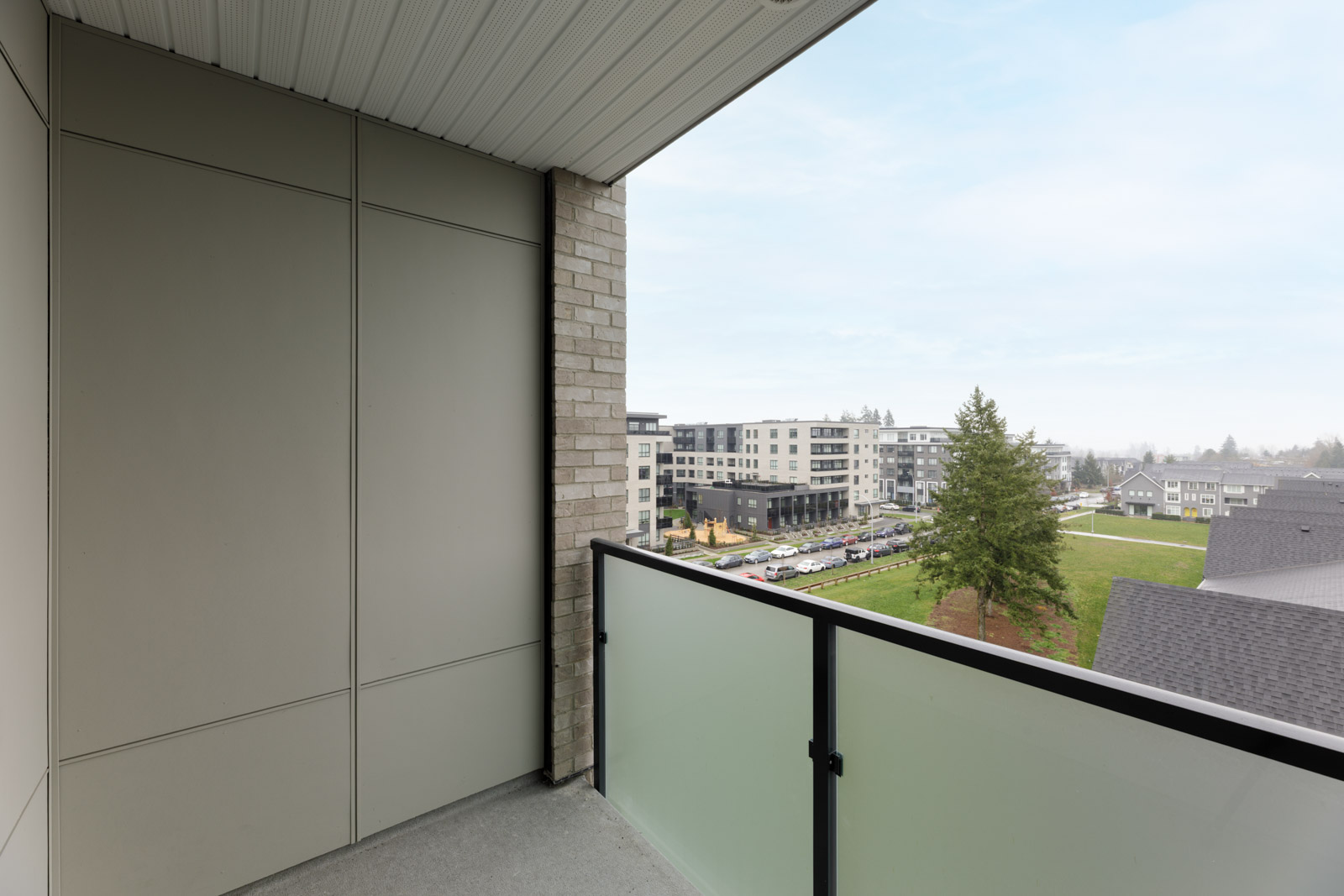 View from a balcony with frosted glass railing overlooking a parking lot, modern apartment buildings, a tree, and grassy areas under a partly cloudy sky.